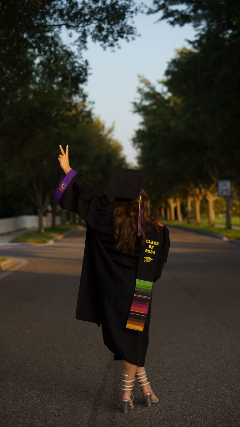 Graduate in cap and gown throwing a peace sign on a tree-lined road